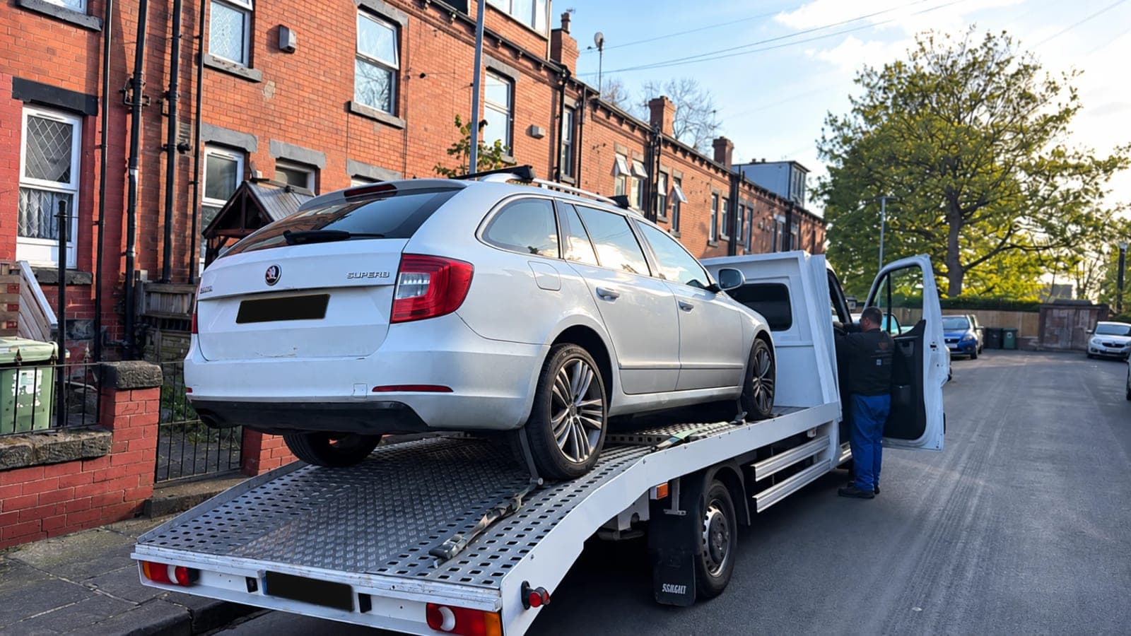 A white Skoda Superb estate winched onto the bed of a white flatbed recovery truck on a terraced street, with the driver standing at the open cab door