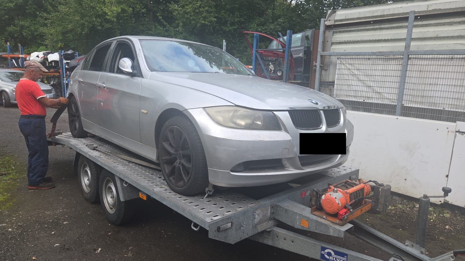 A silver BMW 3 Series on a trailer at the yard, with a member of staff tightening the ratchet strap over a wheel